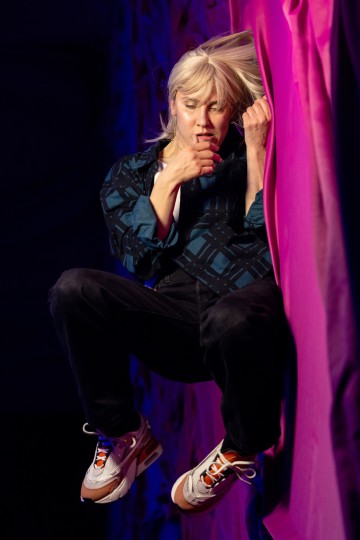 Henkilö, jolla on pitkät vaaleat hiukset, kuvioitu paita, mustat housut ja valkoiset lenkkarit, näyttää leijuvan ilmassa vaaleanpunaisella valaistulla taustalla, silmät kiinni ja toinen käsi lähellä kasvoja.