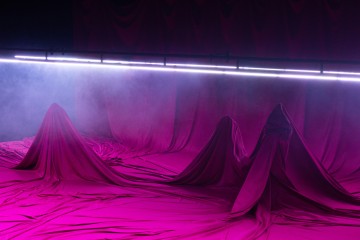 Purppuranpunainen kangas on verhottu useiden lattialla olevien kumpumaisten muotojen päälle, ja taustalla on vaakasuora kaistale kirkasta valkoista valoa ja sumua, mikä luo surrealistisen ja salaperäisen tunnelman.