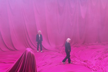 Kolme valkotukkaista hahmoa seisoo ja kävelee vaaleanpunaisella kankaalla päällystetyllä lattialla, ja heidän takanaan roikkuu suuria vaaleanpunaisia verhoja. Yksi hahmo on suurimmaksi osaksi kankaan peitossa, mikä luo salaperäisen tunnelman.