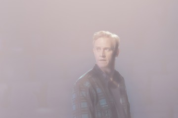 Lyhyet vaaleat hiukset omaava henkilö, jolla on kuvioitu paita päällään, seisoo sumuisessa, sumuisessa valossa ja katsoo hieman sivulle huolestuneena.