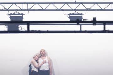 Kaksi valkoisiin tankkitoppeihin pukeutunutta ihmistä seisoo lähekkäin valkoista taustaa vasten. Toinen henkilö painaa päänsä toisen rinnalle, kun toinen katsoo eteenpäin. Heidän yläpuolellaan näkyy näyttämövalaistuslaitteita.