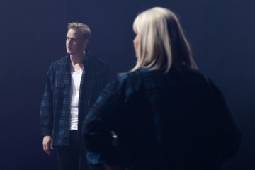 Mies ja nainen seisovat vastakkain hämärässä ympäristössä. Mies näyttää vakavalta, ja hänellä on yllään ruutupaita valkoisen paidan päällä, kun taas nainen näkyy takaapäin ja hänellä on vaaleat hiukset.