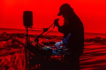 Lakkipäinen henkilö hahmottuu eloisan punaista taustaa vasten, hän käyttää elektronisia musiikkilaitteita ja laulaa tai puhuu mikrofoniin.