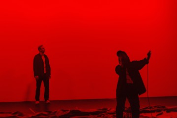Kaksi ihmistä esiintyy lavalla voimakkaassa punaisessa valaistuksessa; toinen seisoo taustalla katsoen ylöspäin, kun taas toinen seisoo etualalla mikrofoni kädessään ja näyttää laulavan tai puhuvan intohimoisesti.