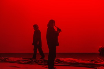 Kaksi ihmistä seisoo lavalla voimakkaan punaisen valon valaisemana; toinen katsoo eteenpäin ja soittaa puhallinsoitinta, kun taas toinen kävelee taustalla. Lattialle on levitetty kangasta.