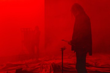 Kaksi ihmistä seisoo punaisen valon ja sumun täyttämässä huoneessa, toinen etualalla pitelemässä mailoja pienen lyömäsoittimen vieressä ja toinen hahmo hämärtyneenä taustalla metallirakenteen vieressä.