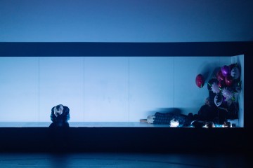 Hämärästi valaistussa näyttämökuvassa yksi henkilö istuu yksin vasemmalla, pää alaspäin, kun taas kolme muuta makaa tai istuu oikealla värikkäiden ilmapallojen joukon kanssa valkoisella taustalla. Tunnelma vaikuttaa synkältä ja pohdiskelevalta.