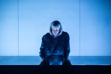 Vaalea peruukki istuu yksin lavalla ja katsoo mietteliäästi alaspäin, sinisen valaistuksen valaisemana, taustan ollessa tavallinen.