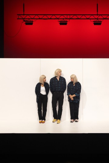 Kolme lyhyen vaaleatukkaista henkilöä, joilla on tummat takit ja mustat housut, seisovat valkoisen seinän edessä lavalla, jossa on punainen tausta ja lavavalot heidän yläpuolellaan. He katsovat toisiaan ja seisovat lähellä toisiaan.