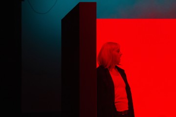Vaaleatukkainen nainen seisoo korkean, tumman suorakulmaisen rakennuksen vieressä, joka on valaistu punaisella ja sinisellä valolla, ja hänen takanaan on kirkkaan punainen tausta. Hänellä on yllään valkoinen paita ja tumma takki.