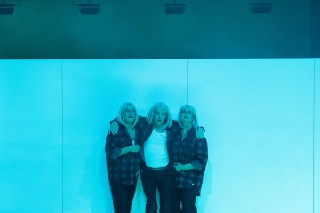 Kolme ihmistä, joilla on samanlaiset vaaleat peruukit ja ruudulliset paidat, seisovat yhdessä sinisessä valaistuksessa, kädet toistensa ympärillä, valkoista taustaa vasten.