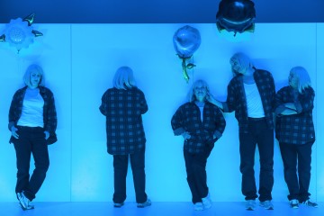 Viisi ihmistä, joilla on samanlaiset hopeiset peruukit ja ruudulliset paidat, seisovat sinisellä valaistulla seinällä. Jotkut katsovat eteenpäin, jotkut kääntyvät poispäin. Heidän lähellään leijuu kaksi metallista ilmapalloa, joista toinen on valkoinen ja toinen musta. Kohtauksessa on surrealistinen, teatraalinen tunnelma.