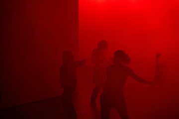 Kolme ihmistä tanssii hämärässä huoneessa, joka on täynnä punaista valoa ja sumua. Heidän siluettinsa ovat osittain peitossa ja liikkeet sumeat, mikä luo dramaattisen ja salaperäisen tunnelman.
