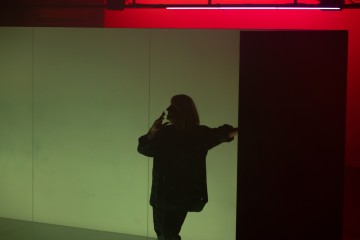 Henkilö seisoo hämärässä tilassa ja pitää puhelinta korvaansa vasten, siluettina taustalla, joka on jaettu pehmeään vihreään ja punaiseen valaistukseen.