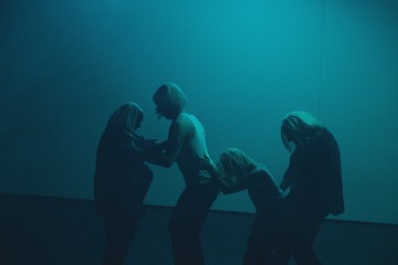 Neljä pitkätukkaista ihmistä seisoo rivissä, kyyristyneinä ja toisistaan kiinni pitäen, siluettina sinistä taustaa vasten.