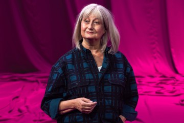 Vanhempi harmaahiuksinen nainen seisoo kirkkaan vaaleanpunaisen taustan edessä, yllään tummansininen ruutupaita. Hänellä on toinen käsi taskussaan ja hän katsoo hieman sivulle lempeä ilme kasvoillaan.