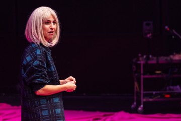 Platinanvaaleat hiukset ja siniruutuinen paita omaava henkilö seisoo sisätiloissa kirkkaan vaaleanpunaisella lattialla ja katsoo hieman sivulle. Tausta on tumma, ja siinä näkyy joitakin laitteita.