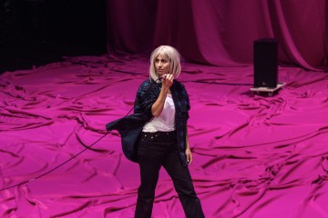 Henkilö, jolla on platinanvaalea polkkatukkainen peruukki, päällään ruutupaita valkoisen t-paidan päällä ja mustat housut, seisoo kirkkaan vaaleanpunaisella kankaalla päällystetyllä näyttämöllä, pitelee narua ja katsoo sivulle.