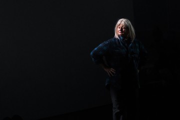 Vanhempi harmaahiuksinen nainen seisoo kädet lanteilla valokeilassa, yllään siniruutuinen paita ja tummat housut, tummaa, varjoista taustaa vasten.