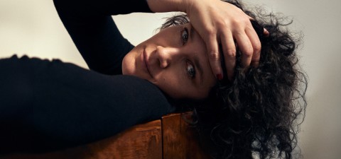 Tummahiuksinen, kihara henkilö, jolla on musta toppi, makaa sivuttain puupinnalla ja katsoo kameraan. Toinen käsi lepää hänen otsallaan, peittäen osittain hänen kasvonsa. Tausta on tavallinen ja pehmeästi valaistu.