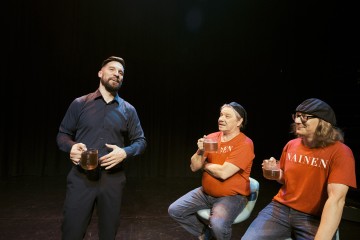 Lavalla kolme miestä mukeja kädessään; yksi mustapukuinen seisoo ja puhuu, kun taas kaksi punapaitaista ja -hattuista istuu ja kuuntelee, taustalla tumma verho.