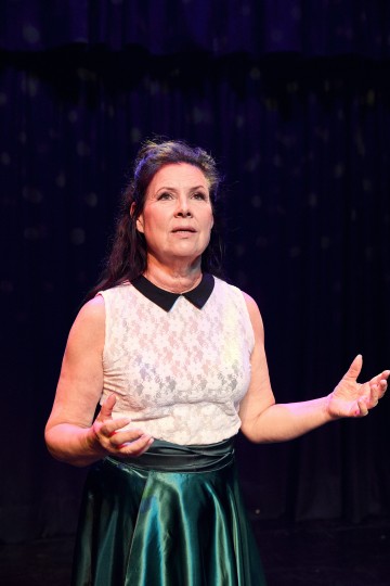 Pitkät tummat hiukset omaava nainen, jolla on yllään valkoinen hihaton pitsitoppi ja vihreä satiinihame, seisoo näyttämöllä dramaattisessa valaistuksessa ja elehtii ilmeikkäästi molemmilla käsillään tummaa verhotaustaa vasten.