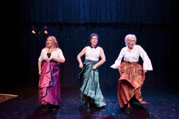 Lavalla kolme naista, joilla on värikkäät hameet ja puserot, tanssivat dramaattista tanssia ja nostavat hameensa ylös, kun he astuvat eteenpäin. Taustalla on pimeä näyttämövalo, joka luo teatterillisen tunnelman.