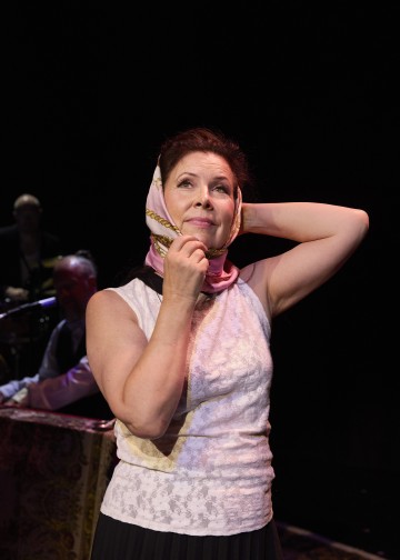 Nainen, jolla on valkoinen hihaton toppi ja vaaleanpunainen huivi päänsä ympärille sidottuna, seisoo lavalla ja katsoo mietteliäästi ylöspäin toinen käsi huivia säätäen. Taustalla on hämärästi valaistu henkilö, joka istuu pianon ääressä.