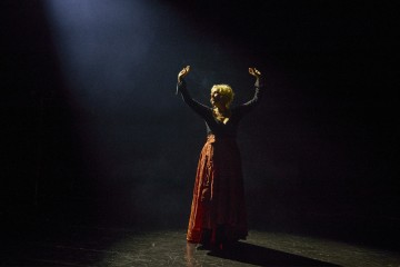 Pitkään tummaan mekkoon pukeutunut henkilö seisoo yksin hämärästi valaistulla näyttämöllä ja kohottaa molemmat kätensä ylöspäin, kun yksittäinen valonheitin loistaa vasemmasta yläkulmasta.