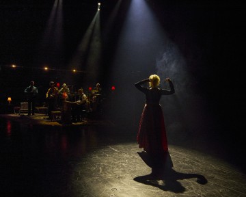 Esiintyjä seisoo valonheittimessä pimeällä näyttämöllä kamerasta poispäin, ja taustalla on valaistu live-bändi. Kohtaus on dramaattinen, ja varjot ja valonsäteet luovat tunnelmallisen tunnelman.