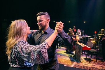 Mies ja nainen tanssivat yhdessä lavalla värikkäiden valojen alla hymyillen toisilleen, kun taustalla soittaa bändi, jossa on näkyviä soittimia, kuten koskettimet, rummut ja kontrabasso.