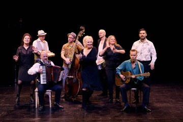 Yhdeksän muusikon ryhmä poseeraa lavalla soittimiensa, kuten harmonikan, kitaran, kontrabasson ja rumpujen kanssa. He ovat pukeutuneet puoliviralliseen tai taiteelliseen asuun ja näyttävät iloisilta lavan valaistuksessa mustaa taustaa vasten.