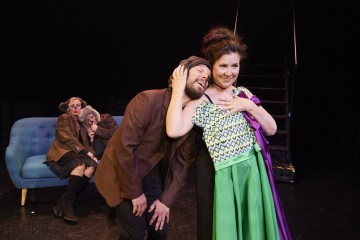 Kaksi näyttelijää poseeraa iloisesti keskellä näyttämöä, ja mies nojaa päänsä naisen olkapäähän. Taustalla kaksi vanhempaa naista istuu sinisellä sohvalla, toinen nojaa päätään toisen olkapäälle. Kaikilla on teatteripuvut.