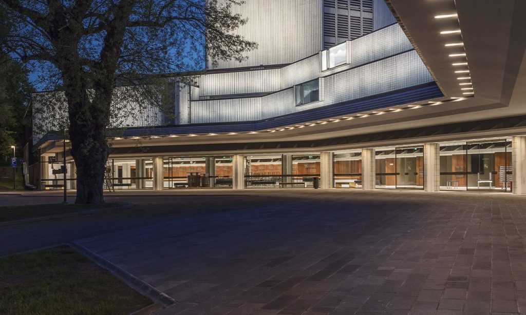 Moderni rakennus, jossa on suuret ikkunat ja geometrinen muotoilu, valaistu hämärässä. Katolla on valorivejä, ja kivetyn sisäänkäyntialueen vasemmalla puolella näkyy puu.