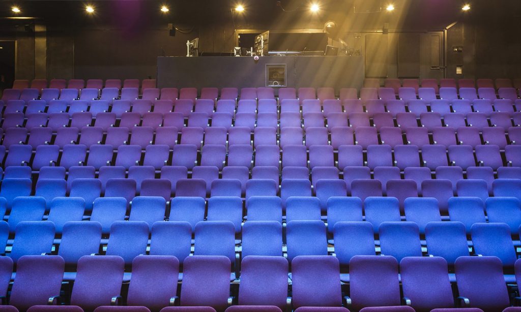 Tyhjien violettien ja sinisten teatteripenkkien rivistöistä on edessä takana oleva ohjauskoppi, jota valaisee näyttämön yläpuolella olevat valot muuten hämärässä salissa.