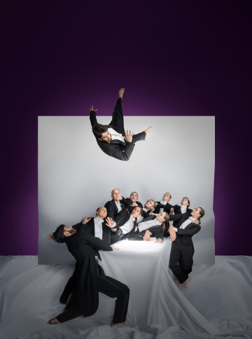 Joukko smokkeihin pukeutuneita ihmisiä poseeraa dramaattisesti valkoista taustaa vasten, ja yksi henkilö on ilmassa ryhmän yläpuolella ikään kuin heitettynä, syvän violettia taustaa vasten.