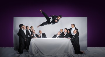 Kaksitoista mustiin pukuihin pukeutunutta ihmistä seisoo ja istuu valkoisen pöydän ympärillä, ja yksi henkilö hyppää dramaattisesti ryhmän yli valkoista ja violettia taustaa vasten. Kohtaus viittaa Viimeiseen ehtoolliseen.