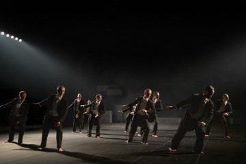 Joukko pukumiehiä, jotka ovat paljain jaloin, tanssivat synkronoitua tanssia hämärästi valaistulla näyttämöllä, ja vasemmalta ylhäältä tuleva valo heittää dramaattisia varjoja näyttämölle.