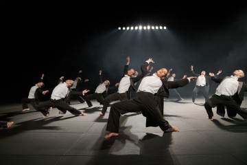 Ryhmä mustiin housuihin ja valkoisiin paitoihin pukeutuneita paljasjalkaisia tanssijoita tekee ilmeikkäitä, synkronoituja liikkeitä hämärästi valaistulla näyttämöllä, jonka taustalla loistavat kirkkaat valot.