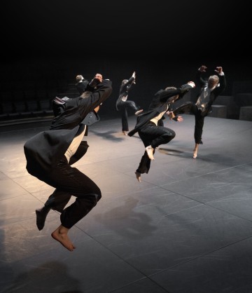 Neljä mustiin pukuihin pukeutunutta tanssijaa loikkii paljain jaloin hämärästi valaistulla näyttämöllä, kädet kohotettuna ja polvet koukussa, ja taustalla näkyy tyhjiä yleisöpaikkoja.