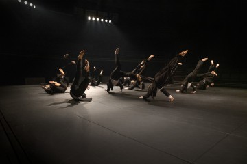 Ryhmä tummiin vaatteisiin pukeutuneita tanssijoita tekee synkronoituja liikkeitä hämärästi valaistulla näyttämöllä, monet heistä tasapainoilevat hartioillaan ja nostavat jalkojaan ilmaan dramaattisten valonheittimien alla.