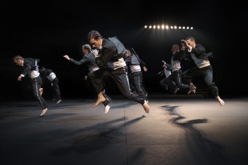 Ryhmä mustavalkoisiin asuihin pukeutuneita paljasjalkaisia tanssijoita loikkii yksimielisesti hämärästi valaistulla näyttämöllä, jonka voimakas ylävalaistus heittää dramaattisia varjoja lattialle.