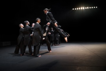 Ryhmä mustiin pukuihin pukeutuneita tanssijoita esiintyy lavalla; yksi tanssija hyppää dramaattisesti ilmaan, kun taas muut seisovat yhdessä lavavalojen valaisemina tummaa taustaa vasten.