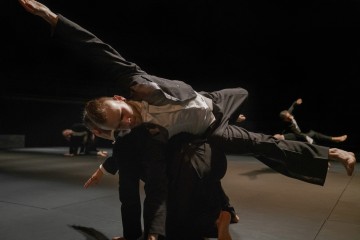 Pukuun pukeutunut tanssija tasapainoilee vaakatasossa toisen esiintyjän selässä kädet ojennettuina nykytanssiesityksen aikana hämärästi valaistulla näyttämöllä. Taustalla näkyy muita tanssijoita.