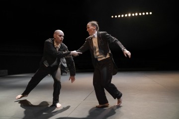 Kaksi paljasjalkaista esiintyjää smokissa tanssii hämärästi valaistulla lavalla. Toinen vetää toista käsivarresta eteenpäin, kun näyttämön valot luovat dramaattisia varjoja taustalle.