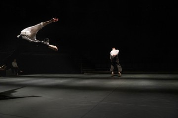 Kaksi tanssijaa esiintyy hämärästi valaistulla näyttämöllä; toinen hyppää dramaattisesti keskellä ilmaa vasemmalla, kun taas toinen seisoo poispäin oikealla. Tausta on tumma, ja yleisön istumapaikkoja on vain vähän.