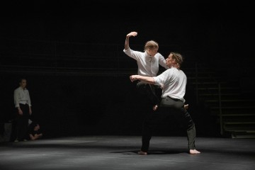 Kaksi valkoisiin paitoihin ja mustiin housuihin pukeutunutta tanssijaa esiintyy hämärästi valaistulla näyttämöllä; toinen nostaa toisen ilmaan. Taustalla näkyy kaksi muuta ihmistä, osittain varjossa portaiden lähellä.