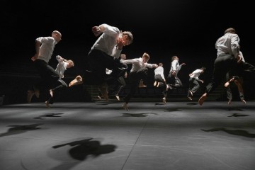 Ryhmä valkoisiin paitoihin ja mustiin housuihin pukeutuneita tanssijoita hyppää ilmaan hämärästi valaistulla näyttämöllä, heittäen dramaattisia varjoja lattialle heidän alapuolellaan.