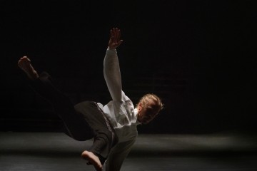 Pitkätukkainen tanssija, jolla on valkoinen paita ja tummat housut, tekee dynaamisen liikkeen hämärästi valaistulla näyttämöllä tasapainoillen yhden käden varassa jalat ojennettuina ja toinen käsi kohotettuna.