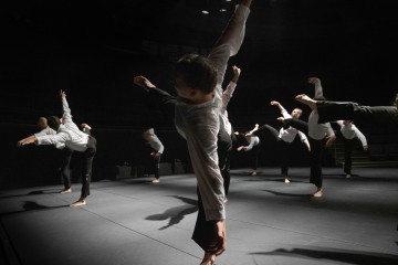 Ryhmä valkoisiin paitoihin ja mustiin housuihin pukeutuneita tanssijoita tekee synkronoituja liikkeitä hämärästi valaistulla näyttämöllä, ja jokainen tanssija tasapainoilee yhdellä jalalla kädet sirosti ojennettuina.